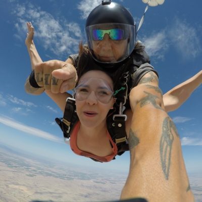 Tandem skydivers mid-air, front person excited, instructor with helmet and tattoos, parachute visible.